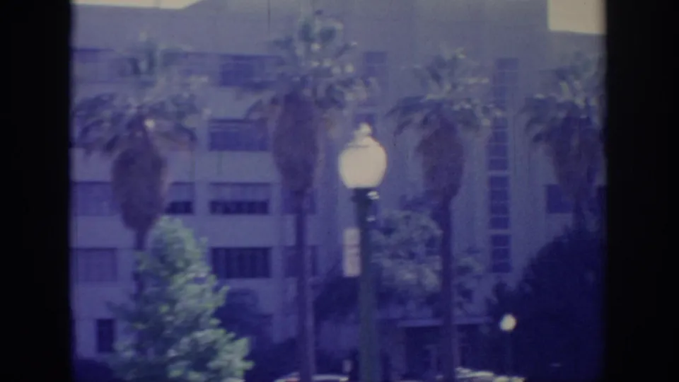 1966 Sacramento home-movie frame showing a multi-story building, palms, greenery, and a white globe street lamp.