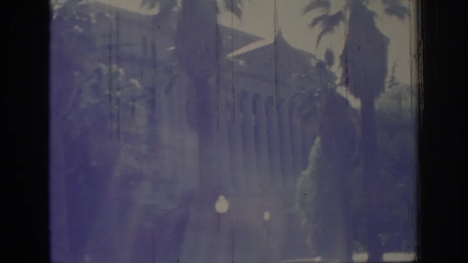 1966 Sacramento home-movie frame showing palms, a partial building view, lamps, and pronounced 8mm gate edges.