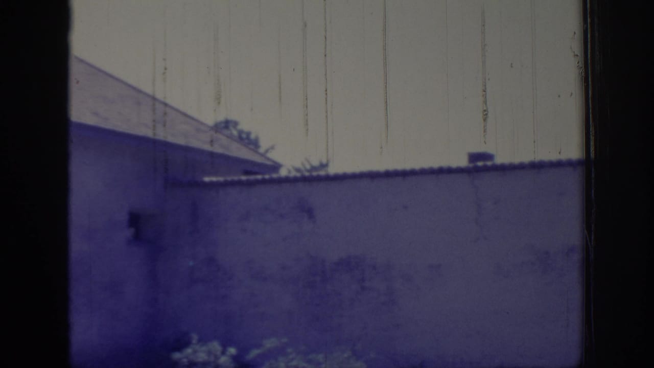 1966 Sacramento home-movie frame showing a light-colored building wall, gabled roof edge, and shrubs viewed through a dark frame.