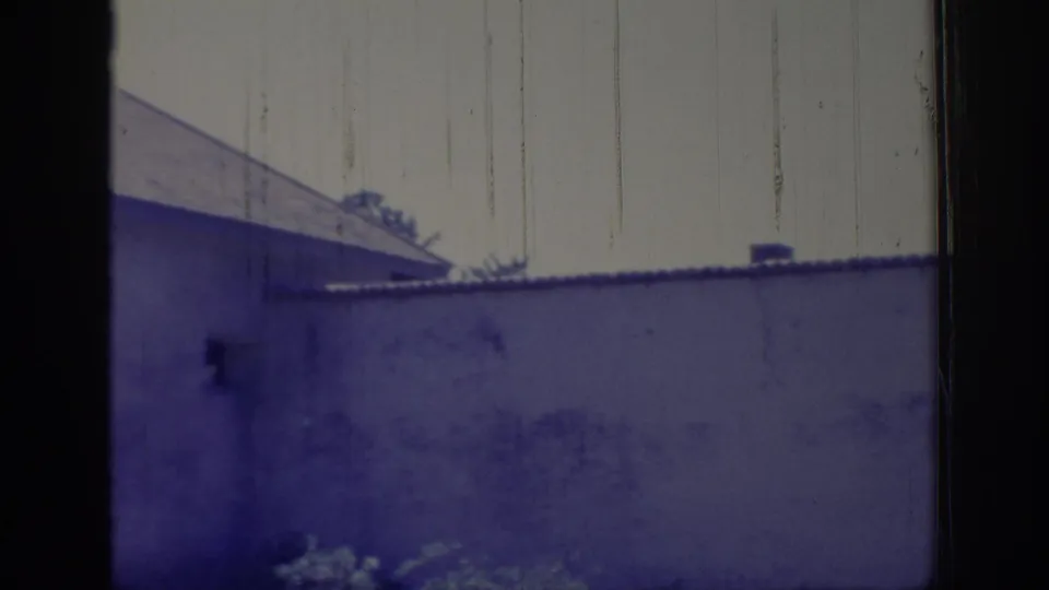 1966 Sacramento home-movie frame showing a light-colored building wall, gabled roof edge, and shrubs viewed through a dark frame.