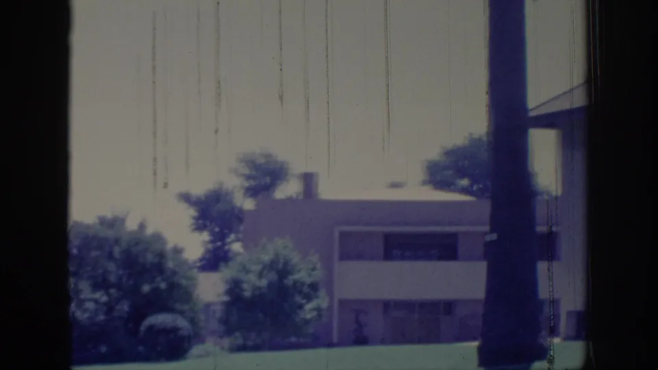 1966 Sacramento home-movie view of a low light-colored building, lawn, shrubs, and a tall trunk partly obscured by dark foreground framing.