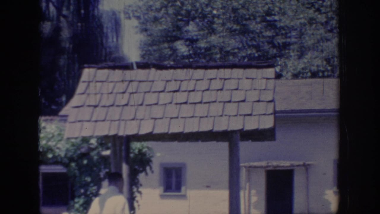 1966 Sacramento frame with shingled arbor roof, yellow cottage wall, open doorway, foliage, and a partly visible adult figure.
