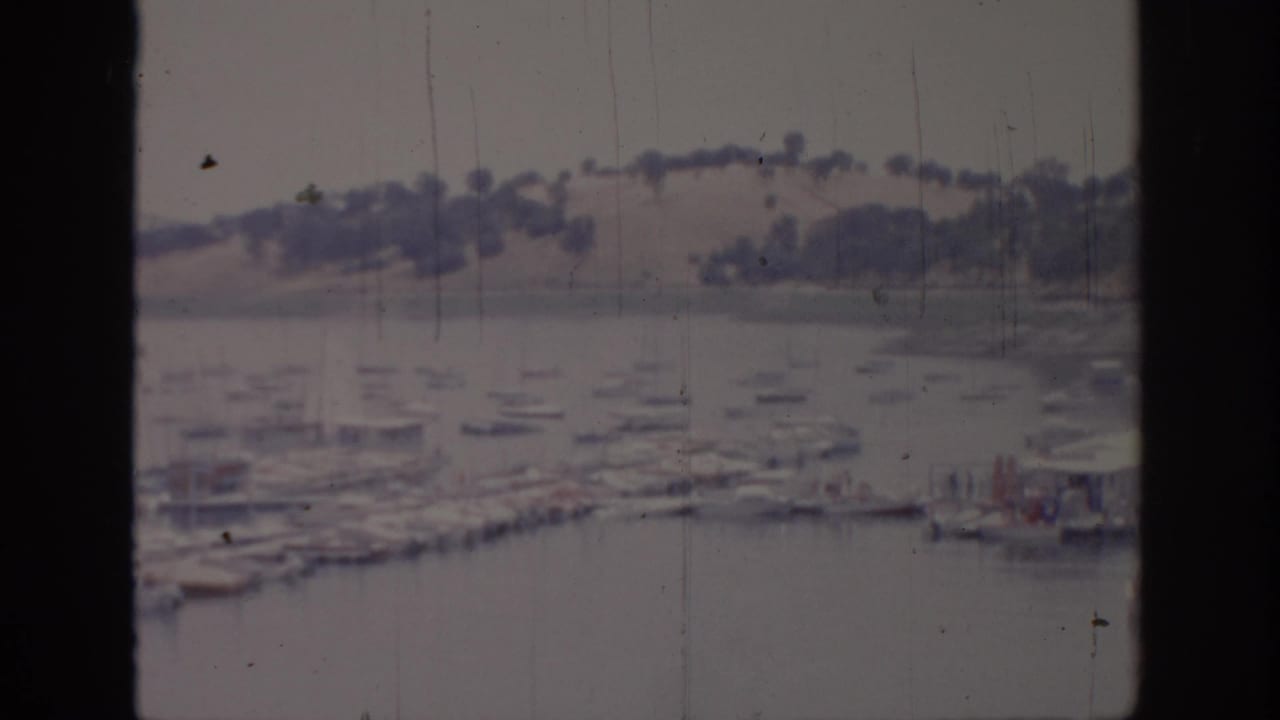 Soft 8mm view of boats docked on calm water with distant hills behind a dark window frame.
