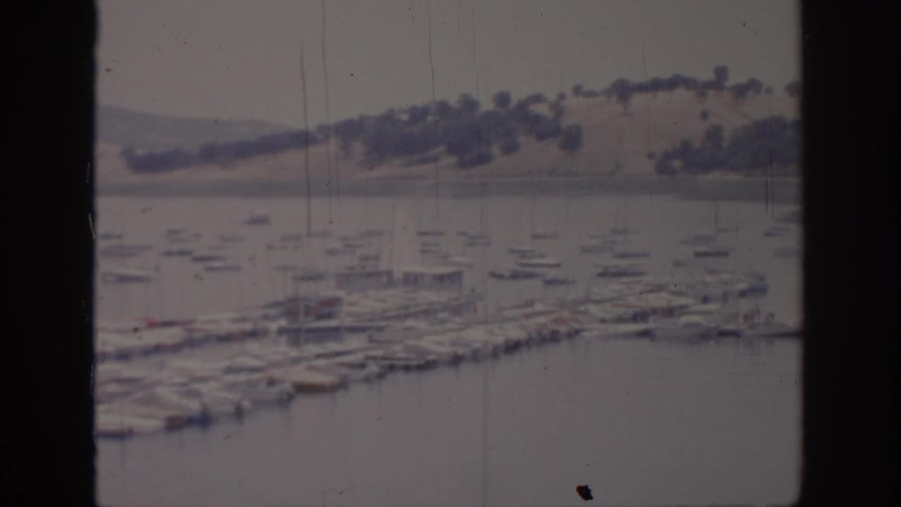 1966 Sacramento-area marina scene with rows of small boats, dock structures, calm water, and low tree-dotted hills beyond.