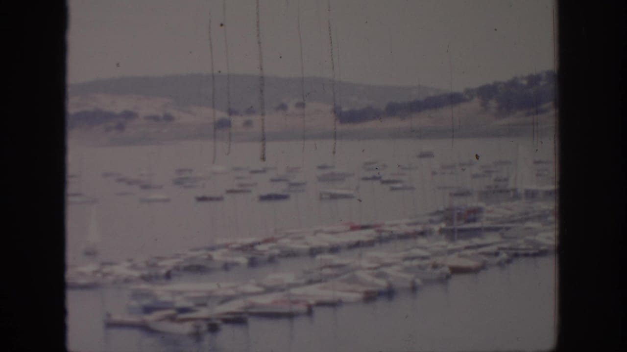 1966 Sacramento-area home-movie frame with docked boats in a marina, still water, distant hills, and vertical window-frame borders.