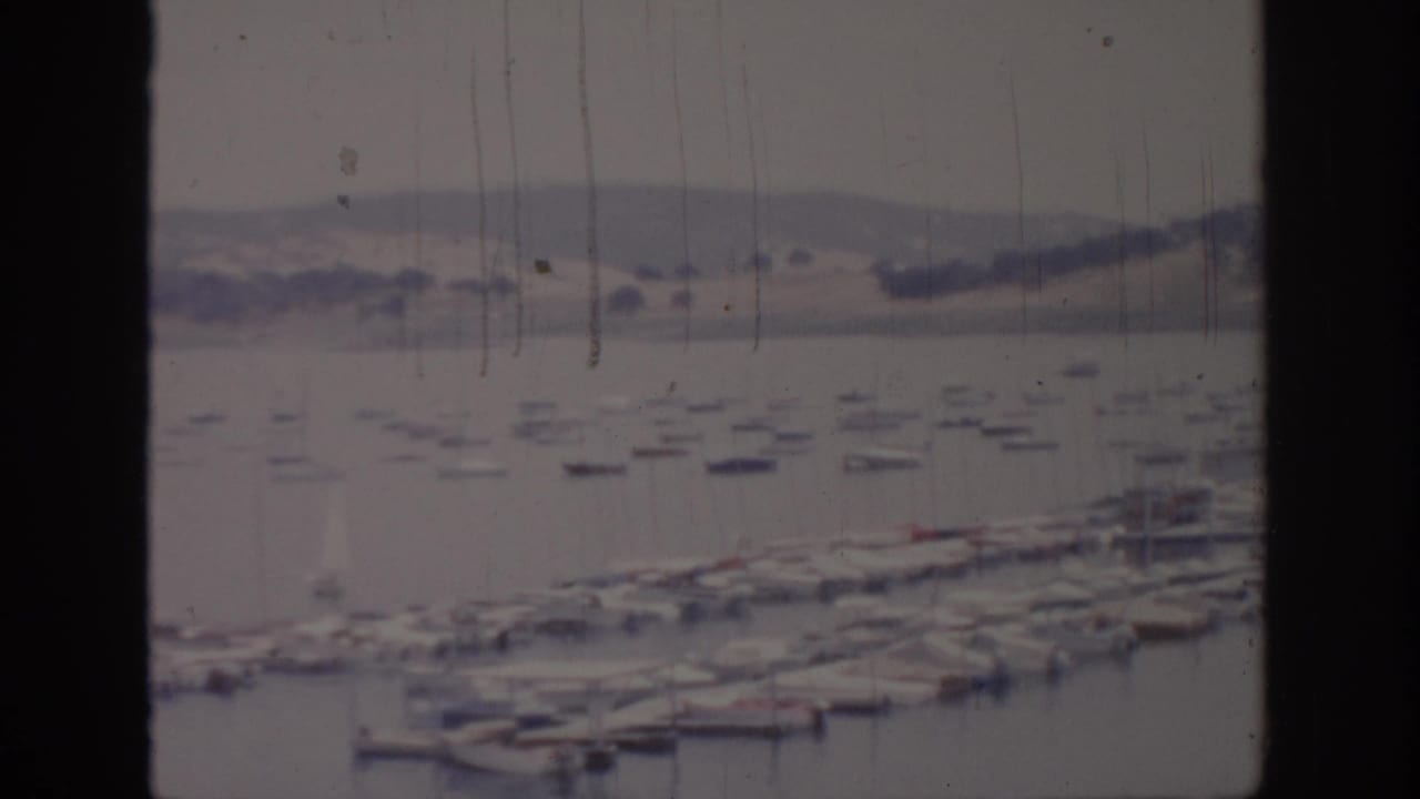 1966 Sacramento harbor view through dark window framing, with small boats on calm water and low hills beyond.