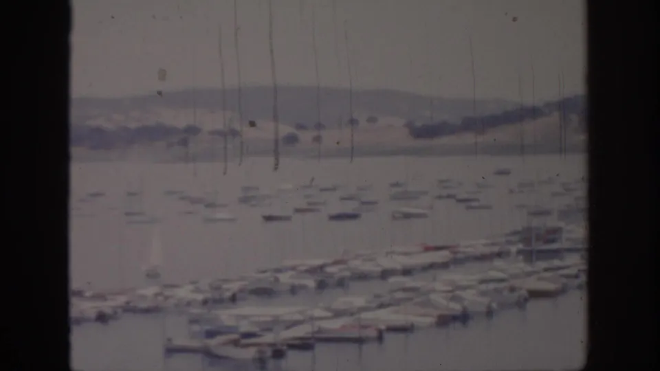 1966 Sacramento harbor view through dark window framing, with small boats on calm water and low hills beyond.