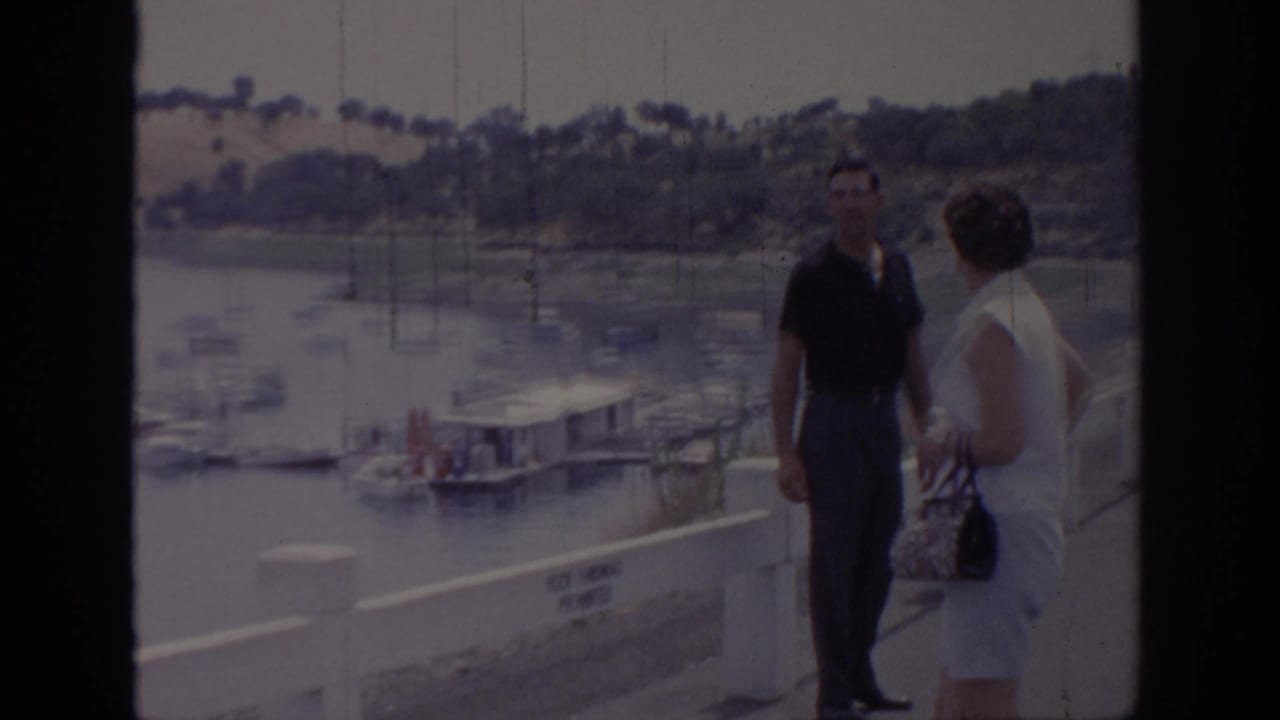 Two adult tourists stand beside a white pier rail at a Sacramento marina, with docked boats and hillside visible behind them.