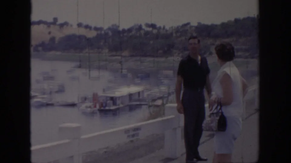 Two adult tourists stand beside a white pier rail at a Sacramento marina, with docked boats and hillside visible behind them.