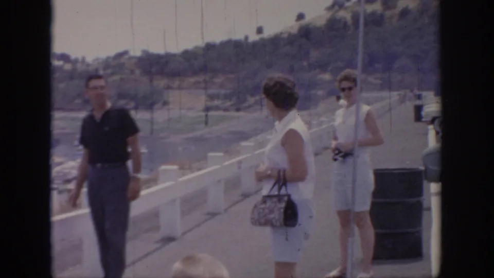Three adult tourists stand on a Sacramento waterfront pier, including a woman with sunglasses and a camera near the marina railing.