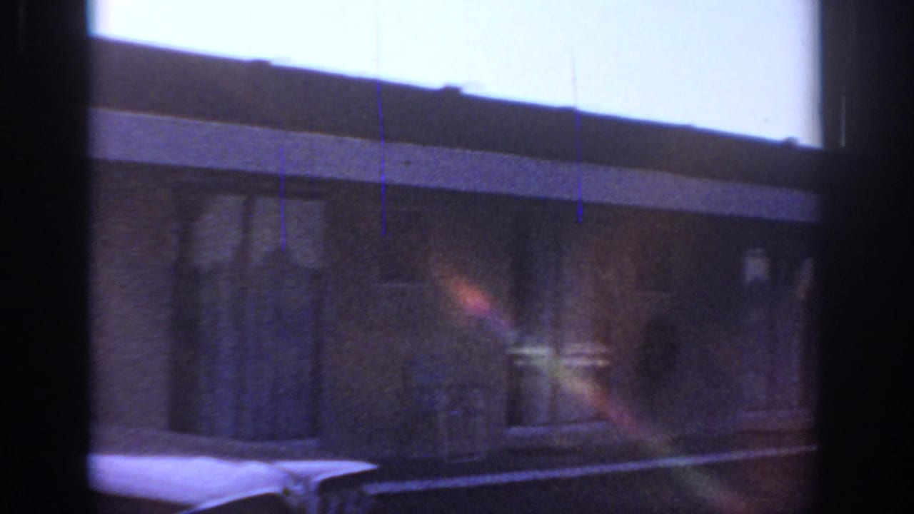 Low building facade and partial car hood seen through a dark window frame.