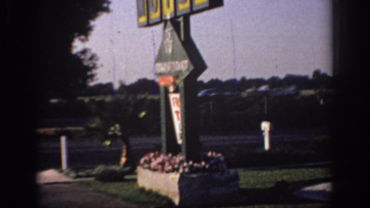 1966 Sacramento 8mm frame showing part of a motel sign, flower planters, roadside lanes, lawn, and distant trees.