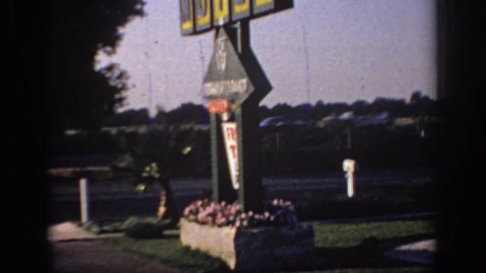 1966 Sacramento 8mm frame showing part of a motel sign, flower planters, roadside lanes, lawn, and distant trees.