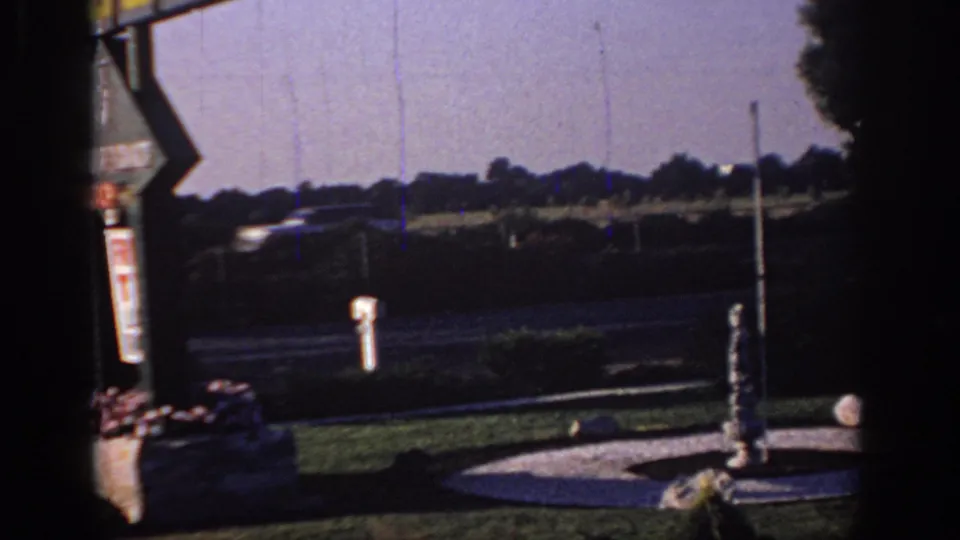 1966 Sacramento motel grounds with a roadside signpost, open lawn, circular fountain area, and distant traffic.