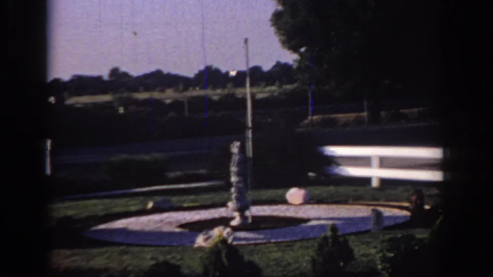 1966 Sacramento motel exterior with a circular fountain, central statue, white fence, lawn, and trees beyond the road.