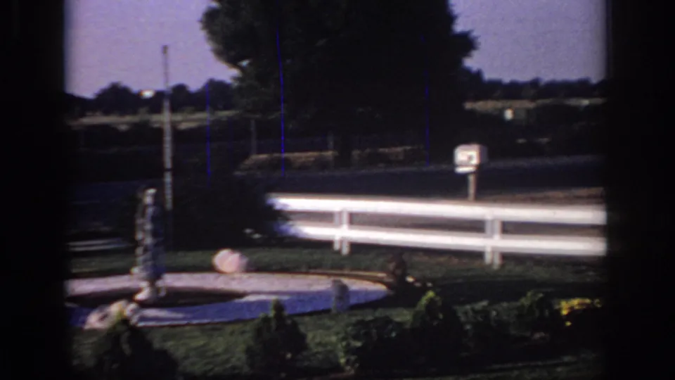 1966 Sacramento 8mm frame showing a motel fountain, central statue, white fence, lawn, shrubs, and roadside background.