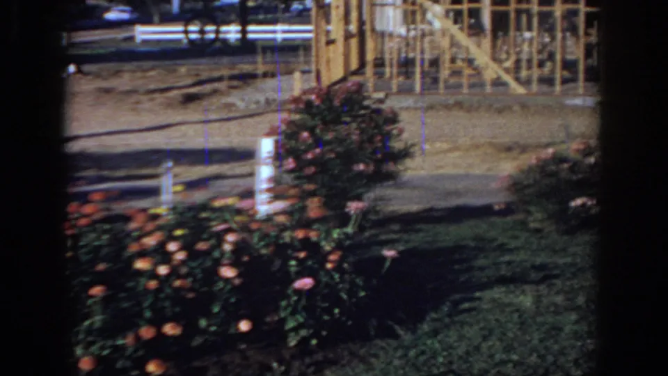 1966 Sacramento home-movie view of roses, lawn, sidewalk, passing cars, and a partial building structure at the right edge.