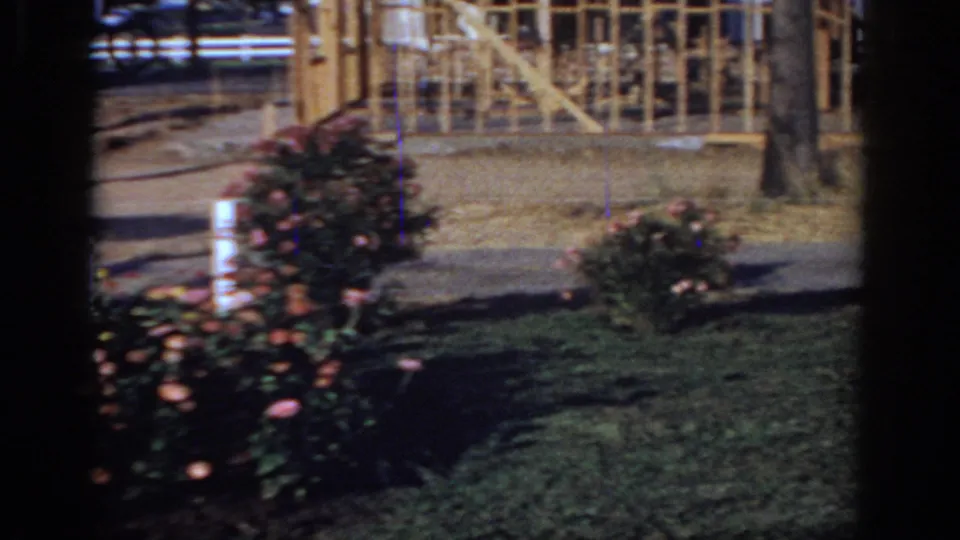 Home-movie view of a Sacramento yard with blooming shrubs, lawn, a tree, and a partially framed structure in the background.