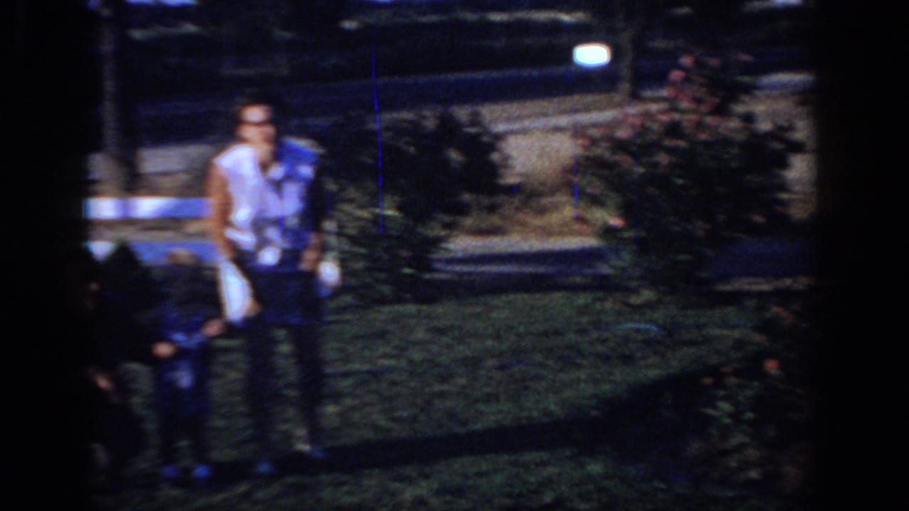 Soft 8mm frame from the Sacramento home movie showing a man near a lawn with a small child or bundle and parked cars in the background.