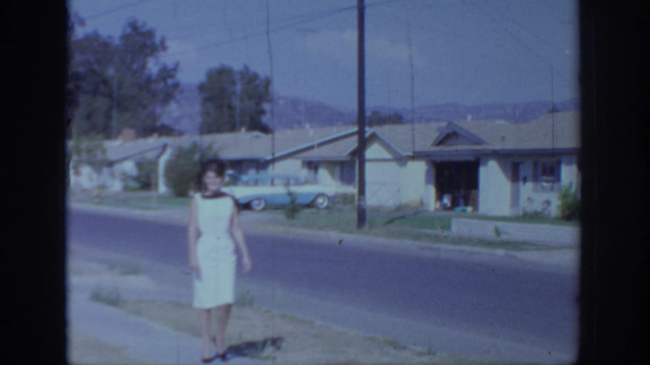 Woman in a white dress standing by a suburban street with houses and a blue car behind her.