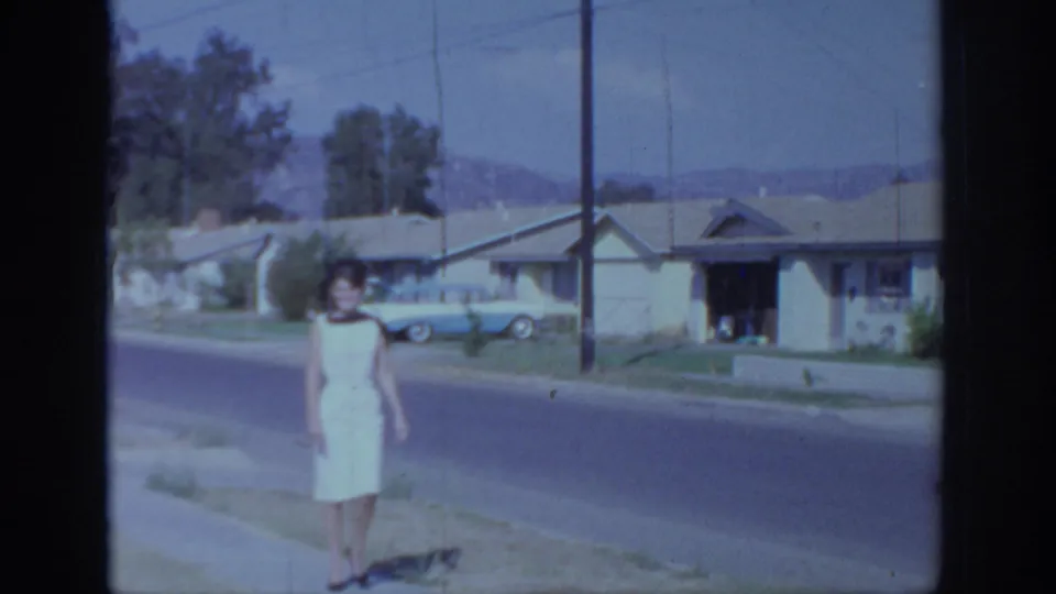 1966 Sacramento frame showing a woman in a white dress near the curb, with single-story homes, utility poles, and a blue car behind her.