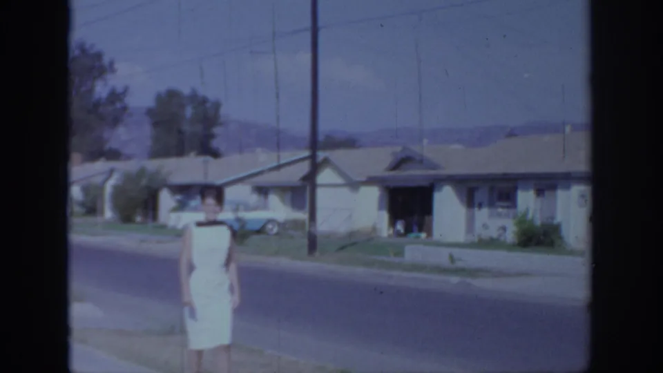 1966 Sacramento home-movie frame with a woman in a white dress walking along a sidewalk past single-story houses and utility poles.