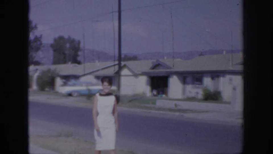1966 Sacramento frame showing the woman in a white dress closer to the camera, standing by the sidewalk in a quiet suburban block.