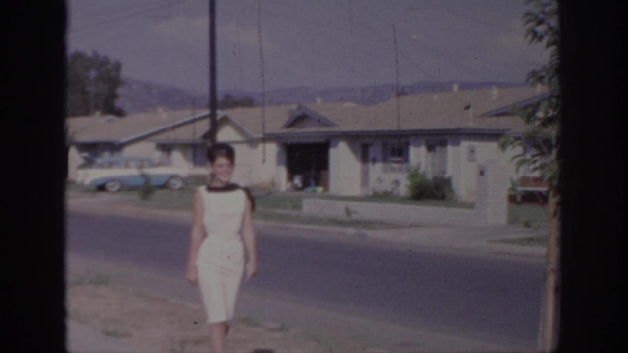 In Sacramento, California, a woman in a white sleeveless dress walks toward the camera on a 1960s suburban street.
