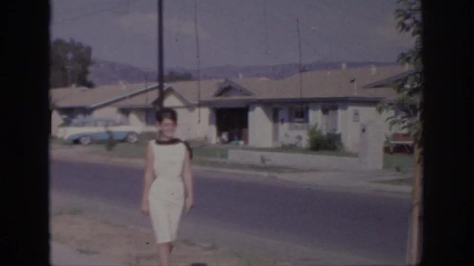In Sacramento, California, a woman in a white sleeveless dress walks toward the camera on a 1960s suburban street.