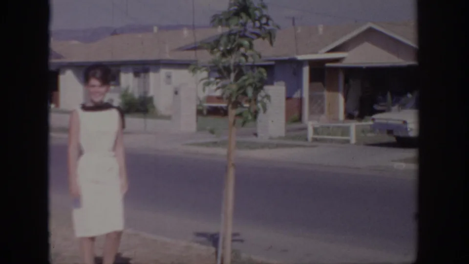 A closer 8mm frame shows the woman smiling as she passes a young tree on a Sacramento residential street.