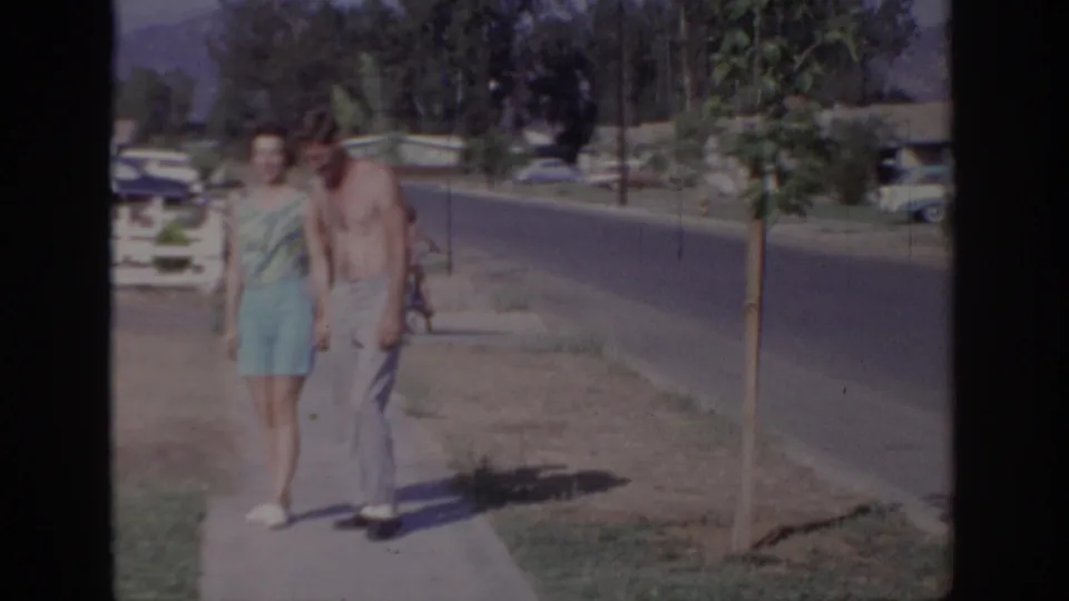 A woman in a turquoise dress and a shirtless man walk toward the camera on a Sacramento sidewalk with cars and houses nearby.