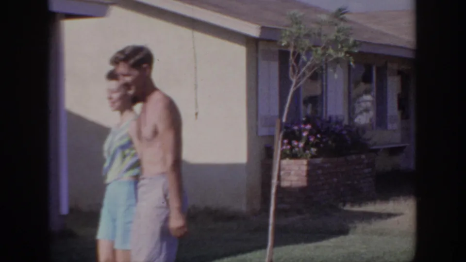 A side view shows the young couple moving past a suburban house, with a lawn, planted flowers, and shaded window behind them.