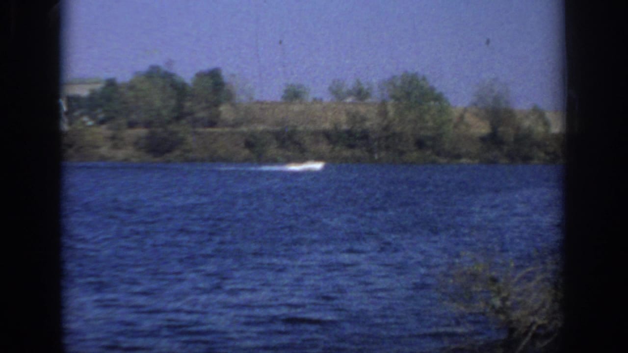 White motorboat wake crossing a blue lake with trees and shoreline in the distance.