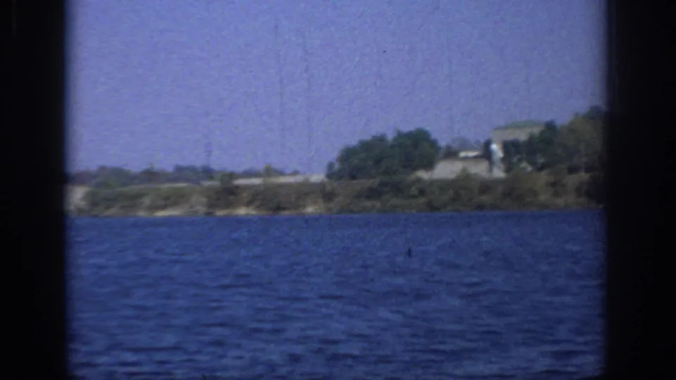 Sacramento 1966 home-movie still of lake water, a tree-lined bank, and distant low structures under a clear sky.