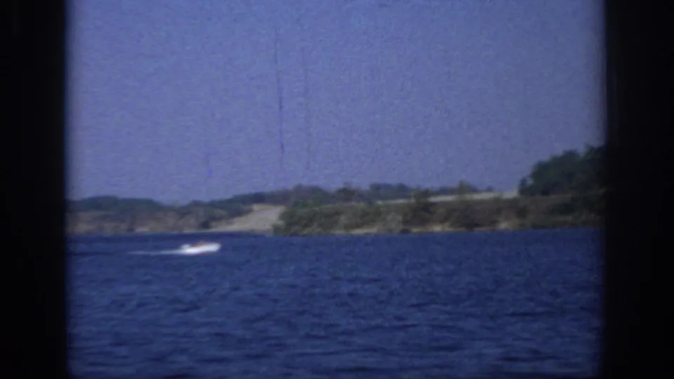 1966 Sacramento lake scene with a small white motorboat wake near the opposite shore and dark 8mm frame edges.