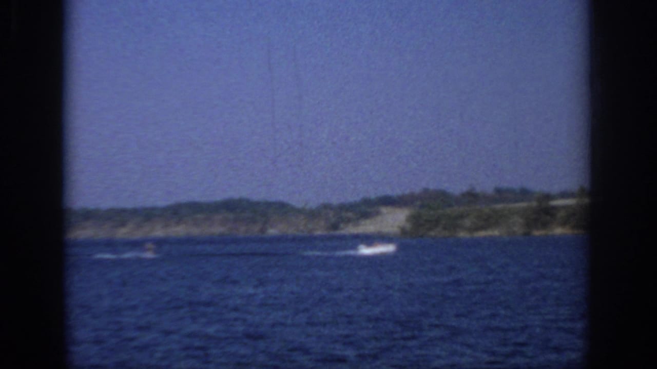Blue lake with distant motorboat wake, wooded shoreline, and dark 8mm frame borders.