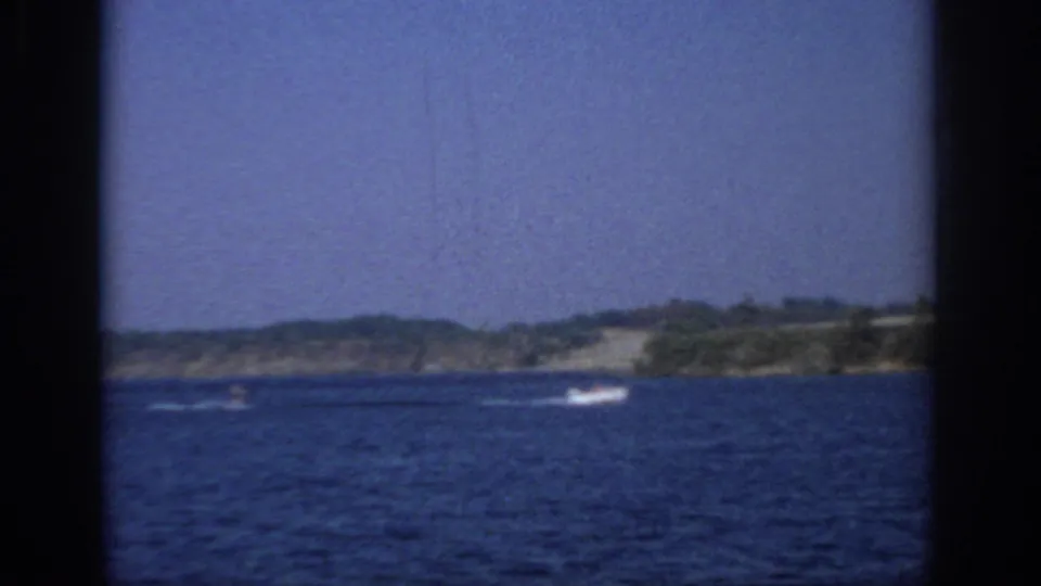 1966 Sacramento-area 8mm frame showing a lake, distant motorboat wake, low wooded shoreline, clear sky, and visible film gate borders.