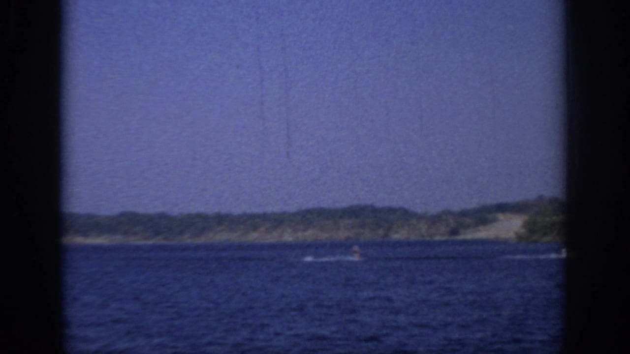 Wide lake view with distant shoreline, small boat activity, blue sky, and film borders.
