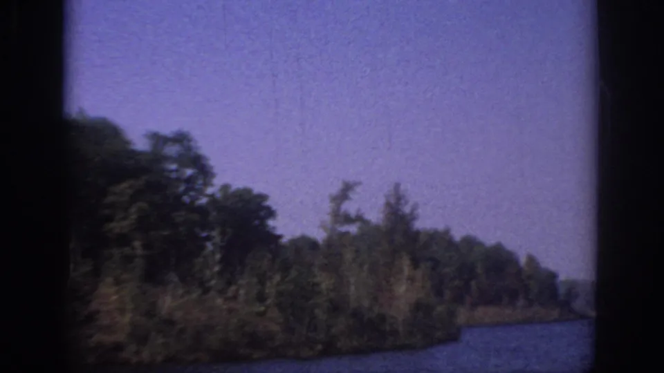 1966 Sacramento-area 8mm frame with a wooded bank, open blue water, soft focus, and dark vertical film borders.