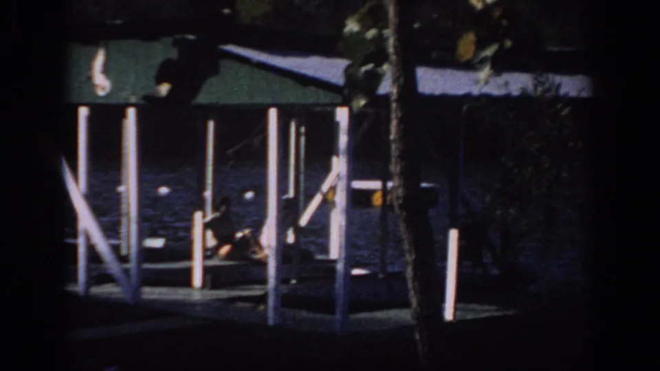 1966 Sacramento-area home-movie frame with a distant person seated on a covered dock above dark lake water.