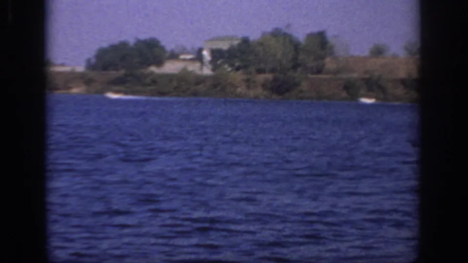 1966 Sacramento-area 8mm frame looking across blue lake water toward a distant tree-lined shoreline and small boats.