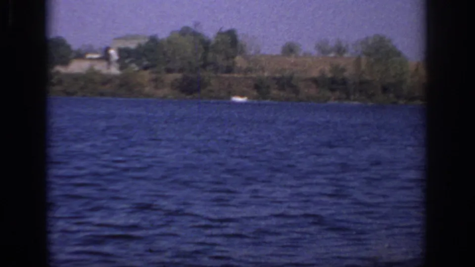 1966 Sacramento-area home-movie frame showing open blue water, distant trees, low shoreline structures, and small boats.