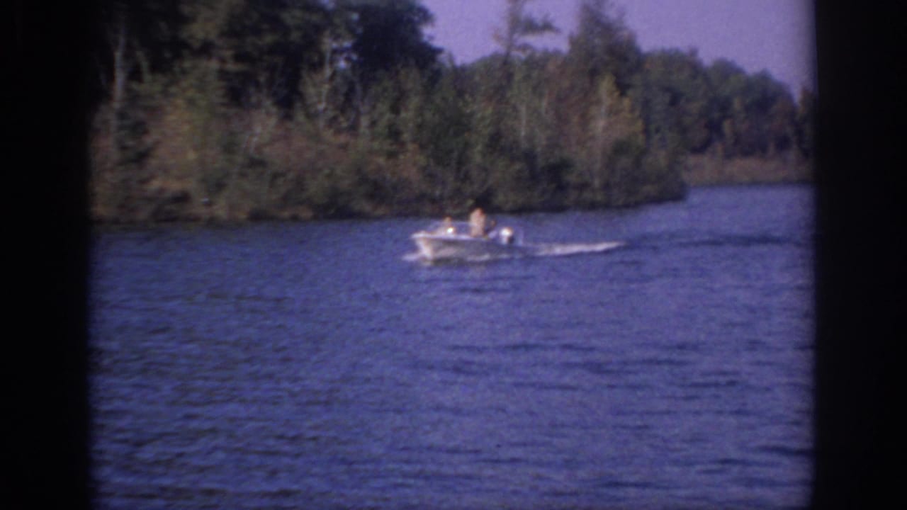 1966 Sacramento frame of a white outboard motorboat cruising on a calm body of water near a tree-lined shore.