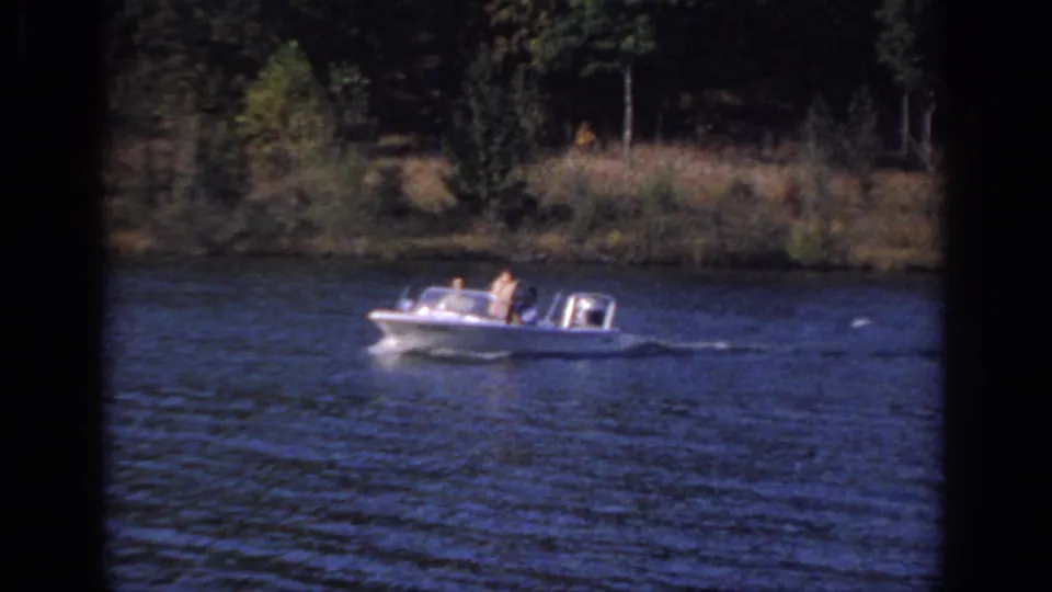 1966 Sacramento home-movie frame showing a white windshield motorboat with passengers passing a wooded bank.