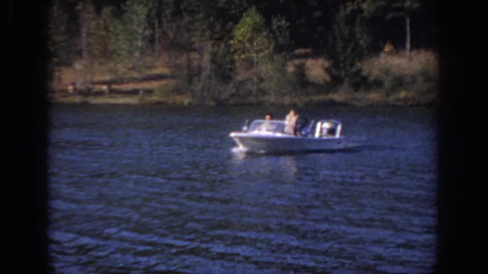 1966 Sacramento frame of a small white powerboat with visible passengers on rippled water near a wooded shoreline.