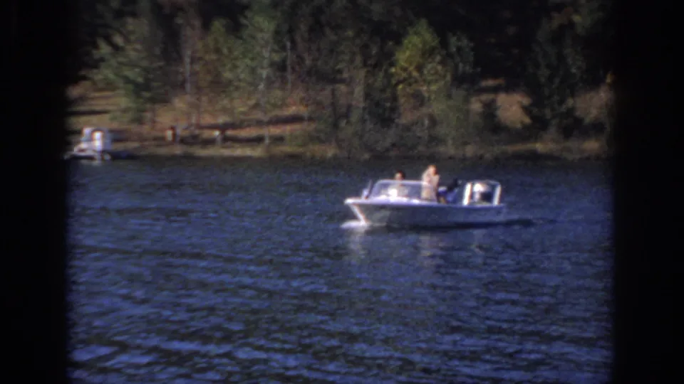 1966 Sacramento-area home-movie frame showing a small motorboat with passengers moving across rippled lake water near a wooded shore.
