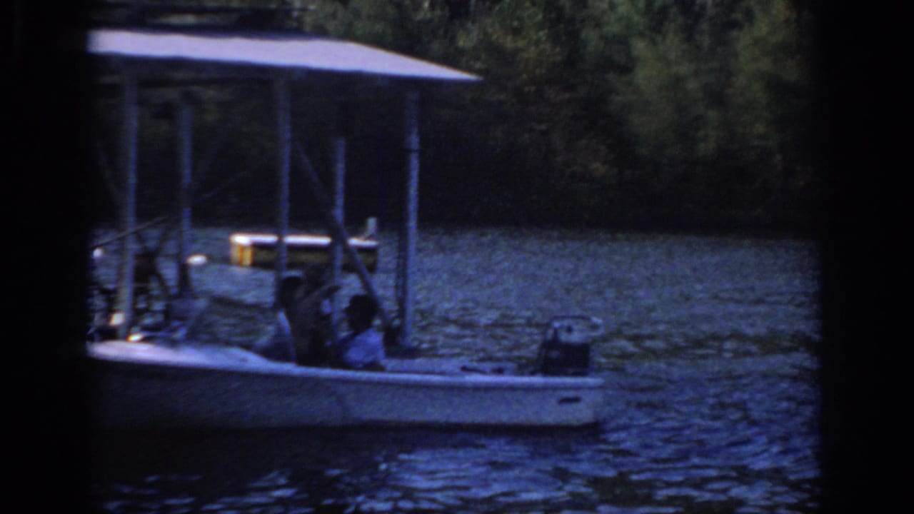 A covered boat or docked craft sits on the lake, with seated figures partly obscured by posts and deep 8mm shadow.