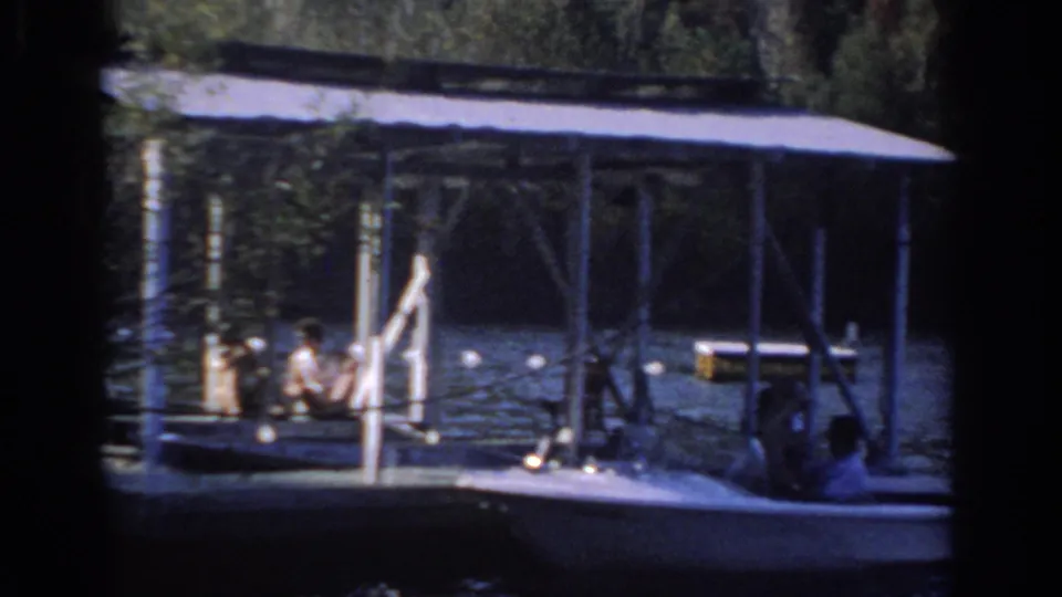 Lake scene with people near a covered boat and dock-like structure, framed by trees and dark 8mm film edges.