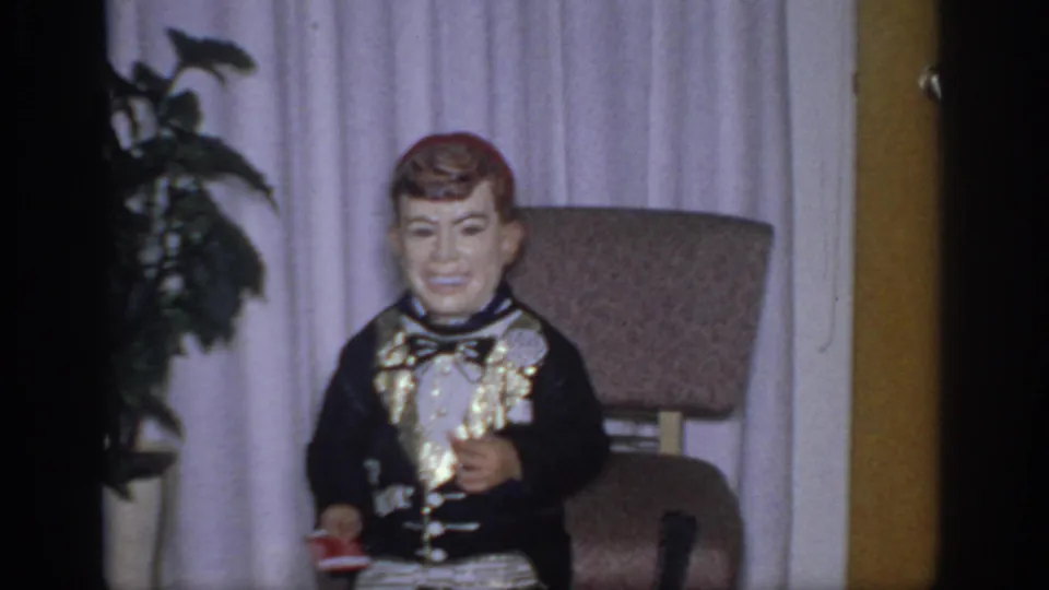 Indoor home-movie frame of a child-sized costumed figure with an oversized head, bow tie, curtains, plant, and upholstered chair.