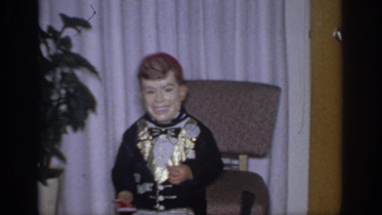 1966 Sacramento home-movie frame showing a painted ventriloquist dummy in a black formal outfit near curtains, a chair, and a houseplant.